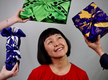 Artist Keiko Kira in the middle surrounded by hands holding gifts wrapped in green, purple and blue Japanese furoshiki