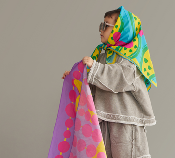 A toddler looking afar, wearing art play silk in dots and friends print as a head scarf and holding one in hide and seek print