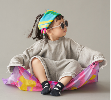 A toddler sitting on top of art play silk in hide and seek print smiling and looking to the right