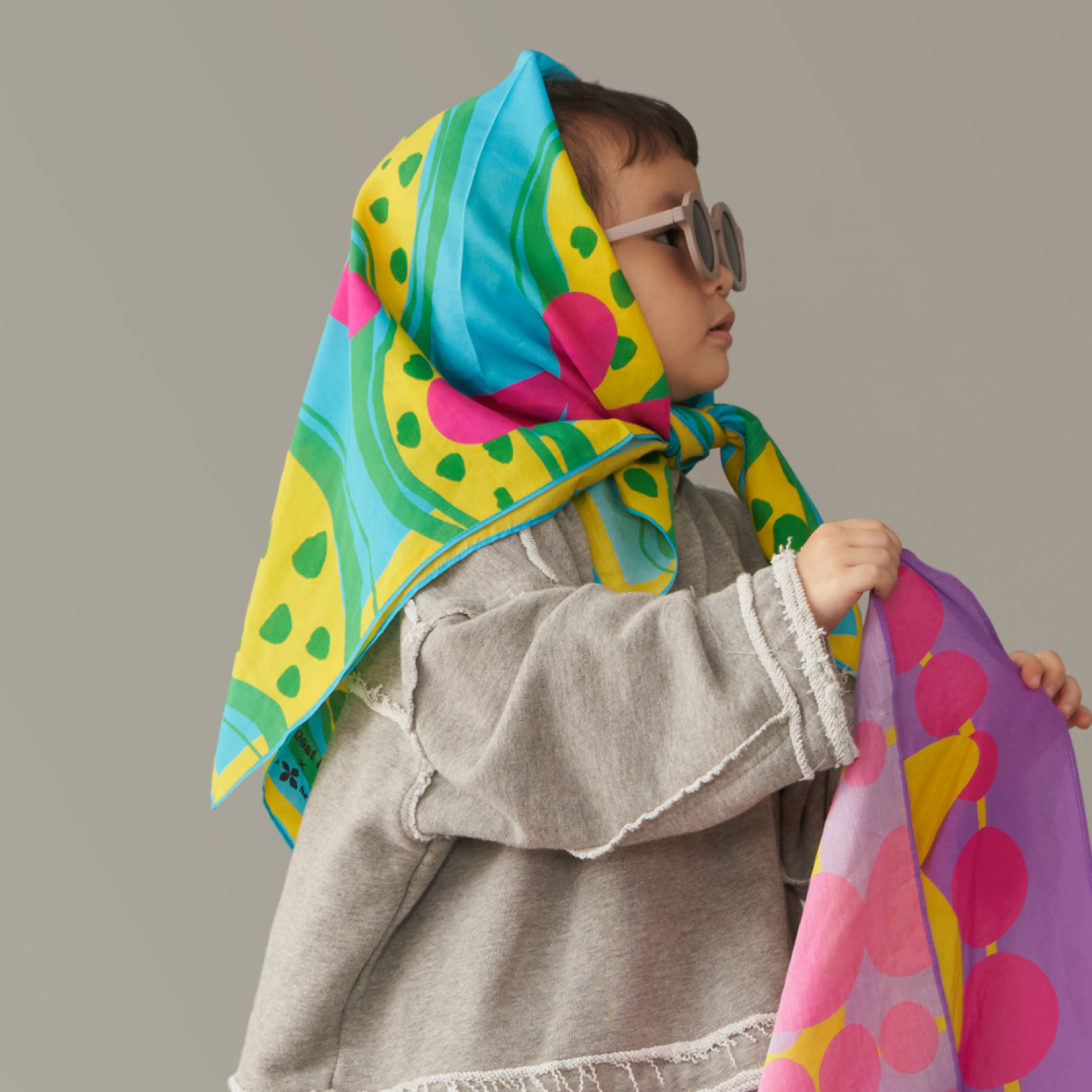 Child wearing art play silk in "dots and friends" print - blue and green design - as a head scarf holding another art play silk