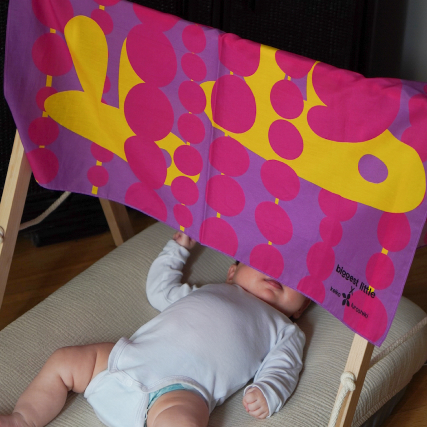 Baby lying on a mat with a colorful art play silk in hide and seek print featuring pink and yellow abstract designs hanging over the baby